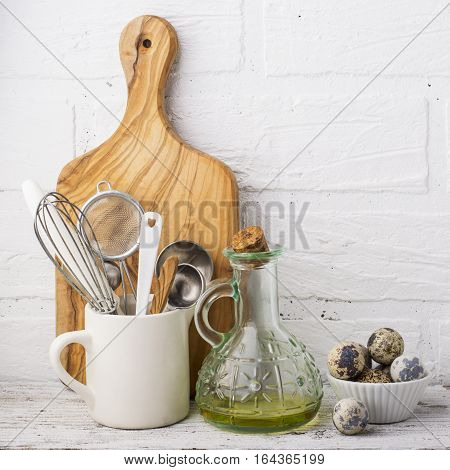 Kitchen Still Life. Olive oil in a jug, quail eggs and kitchen tools for baking, olive cutting board on a wooden shelf in the background of a white brick kitchen wall. selective focus
