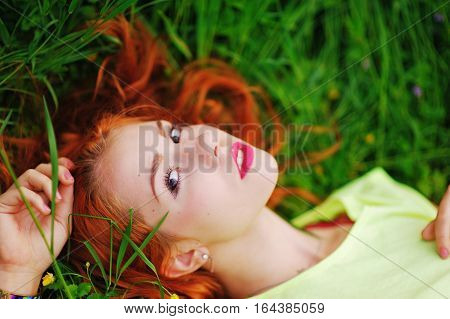 beautiful red-haired girl in a yellow shirt lying on the grass and stares dreamily