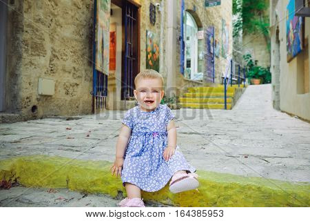 Portrait Of Cute Baby Girl Sitting On A Rock Step Outdoors And Smiling