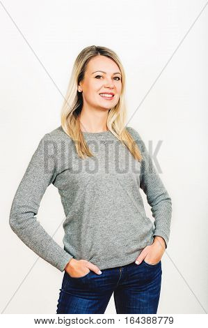 Studio shot of young blond woman wearing grey pullover and denim jeans, hands in pockets