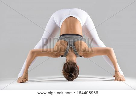 Young attractive woman practicing yoga, standing in Wide Legged Forward Bend exercise, Prasarita Padottanasana pose, working out wearing sportswear, indoor full length, grey studio background
