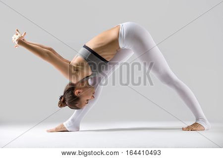 Young attractive woman practicing yoga, standing in One Sided Fold, Pyramid, exercise, Parsvottanasana pose, working out wearing sportswear, indoor full length, isolated against grey studio background