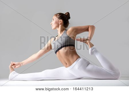 Young attractive woman practicing yoga, sitting in Monkey God, Splits exercise, Hanumanasana pose, working out wearing sportswear, white pants, top, indoor full length, against grey studio background