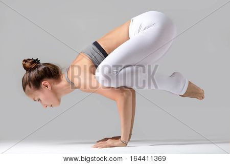 Young attractive woman practicing yoga, standing in Crane exercise, Bakasana pose, working out wearing sportswear, white pants, gray top, indoor full length, isolated against grey studio background