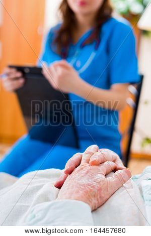 Senior patient's hands folded in bed in residential care home and a nurse in the background examining the patient.