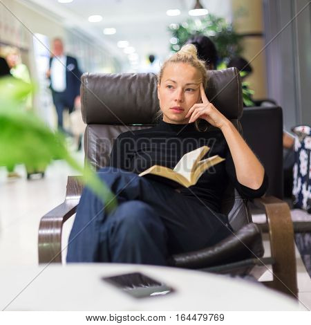 Thougtful woman sitting by wooden table and reading book in public library.