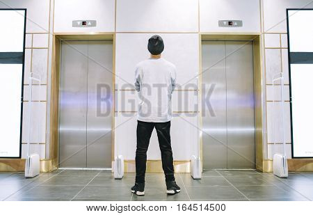 Man in jeans and hat standing between the elevators with crossed arms on his chest. Young guy is waiting for a lift. View from the back