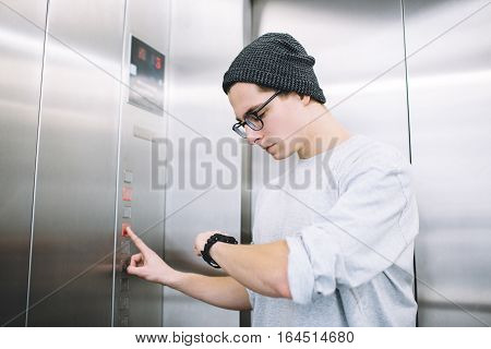 Young stylish guy standing in elevator and pressing button to go up. Serious boy is checking his time for not to be late for meeting