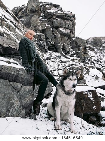Girl with dog Malamute among rocks in winter. They sit on rocks among scattering of stones and snow.