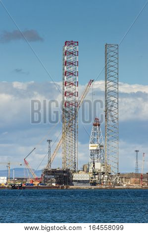 Balnapaling Scotland - June 3 2012: Galaxi Z petroleum and national gas drill platform under construction in the harbor. Tall frames and cranes. Heavy metal contraption. North Sea in front under blue cloudy sky.