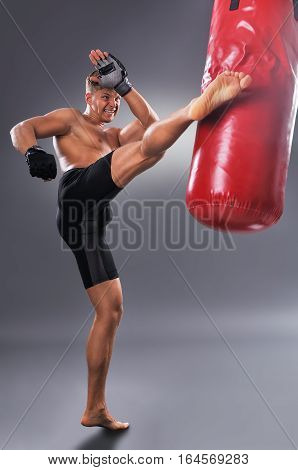 Muscular Fighter Practicing Some Kicks With Punching Bag In Black Boxing Gloves. Boxing On Gray Back