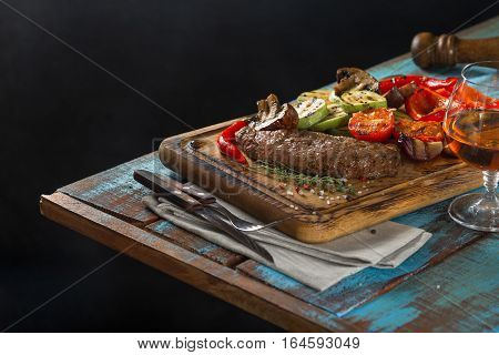 Steak grilled with grilled vegetables and glass of brandy on a dark background