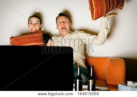 a couple is sitting on a couch and watching a VERY scary movie scene on tv. the shock is so great that pillows and popcorn are flying around.