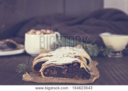 Slice of fresh baked homemade strudel with poppy seeds and sugar powder on a black background and fir branches. Cocoa with murshmelou. Christmas winter still life