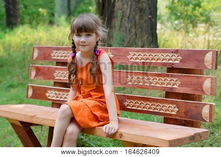 Happy cute little girl in orange sits on wooden beanch at summer day