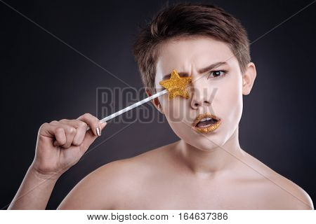 Emotionaly confused. Pleasant puzzled woman holding lollipop and looking at you while standing against black background