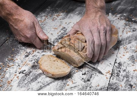 Baking and cooking concept background. Hands of baker cutting bread loaf with knife on rustic wooden table sprinkled with flour. Stained dirty hands of baker