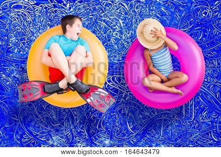 Raucous little boy and his quiet young sister relaxing together on bright colorful plastic rings in the swimming pool enjoying a hot summer day conceptual image on blue painted water background with ripples
