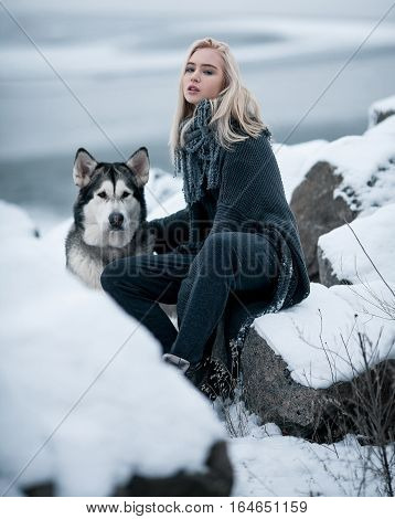 Girl with dog Malamute among rocks in winter. They sit on rocks among stones and snow.