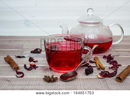 Hibiscus tea in glass teapot, cap with tea, spices and dry flower on wooden background