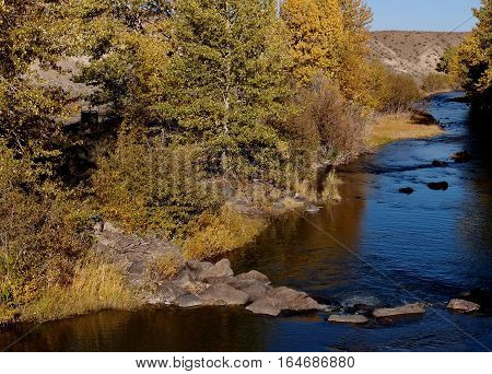 The bright blue waters of the Chewaucan River near Paisly in Southeastern Oregon with colored trees and grasses on its banks as well as a rock outcropping on a sunny fall day.