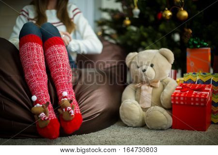 Close up of female legs in red christmas socks with fun fluffy elks. Christmas gifts and teddy bear under decorated pine tree.