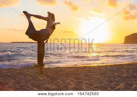 Silhouette of young woman practicing standing bow yoga pose on sandy beach at sunset.