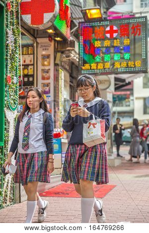 Hong Kong, China - December 6, 2016: asian teens in typical school uniform with mobile phone down the street market Jardine's Crescent, Causaway Bay, on the road full of shops with bright signboards.