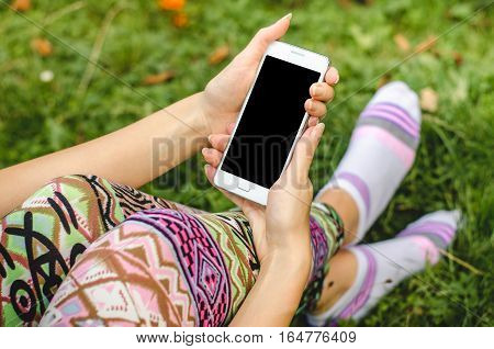 close up white mobile phone in fashionable trendy woman hands on the background of green grass with flowers during an exercise break