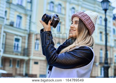 Beautiful girl photographer photographing the old town with retro camera. She is wearing a knitted cap