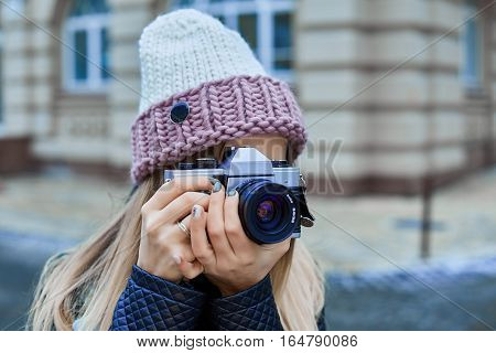 Beautiful girl photographer photographing the old town with retro camera. She is wearing a knitted cap