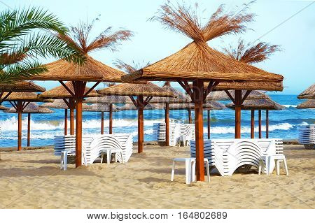 Empty beach in the seaside resort with folded sun loungers and wooden sheds with thatched roof on a background of blue sea waves.