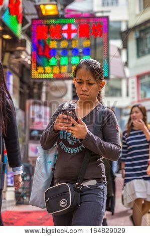 Hong Kong, China - December 6, 2016: asian woman walking and reading her mobile phone in street market Jardine's Crescent, Causaway Bay, shopping district, full of street shops with shining signboards