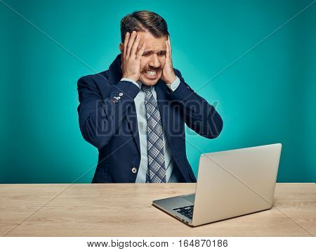 The sad and puzzled young man in a business suit working on laptop at desk on blue studio background