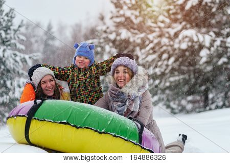 Two women and little child hugging and laughing on snow tubing. Group of caucasian adults and kid having fun outdoors. Winter vacation.