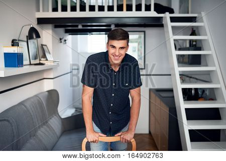 Portrait of a handsome young man smiling and leaning on a chair while standing in his modern loft apartment