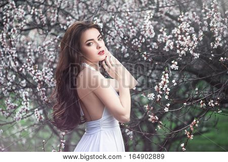 Sensual young woman standing in white dress at blossoming tree in the garden. Beauty of woman and nature consept. Portrait of beautiful model with curly brown hair posing and looking at camera.
