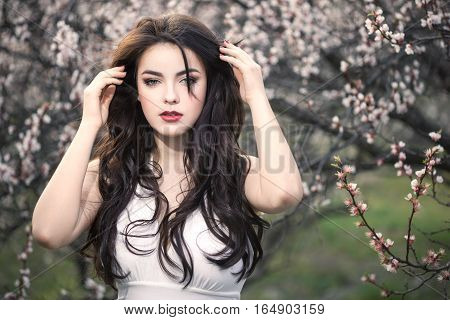 Sensual young woman standing in white dress at blossoming tree in the garden. Beauty of woman and nature consept. Portrait of beautiful model with curly brown hair posing and looking at camera.