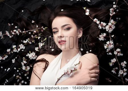 Spring beauty consept. Portrait of a fashionable model with curly brown hair sexy red lips lying on black background with spring trees blossoming branches and petals background. Studio portrait shot of gorgeous woman in white dress with perfect make-up se