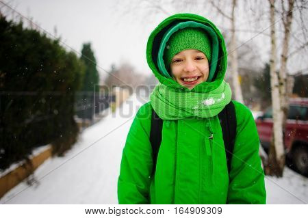 The cheerful teenager goes down the street in winter day. Behind shoulders at him a backpack. The boy is dressed in a bright green ski suit. It is snowing. The fellow smiles from pleasure.