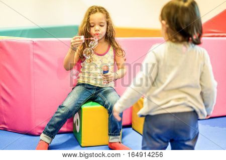 Two little female children blowing soap bubbles in playground nursery school - Young people having fun indoor - Friendship and game concept - Focus on left child face - Warm vivid filter