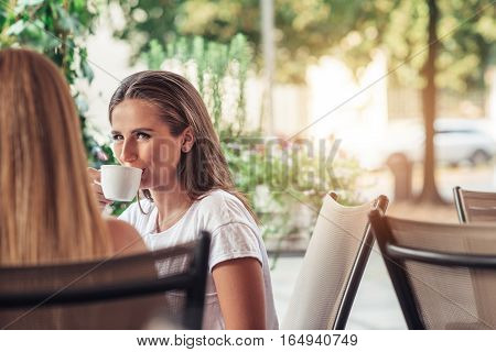 Attractive young brunette woman drinking coffee and talking to a friend while sitting outside at a cafe table