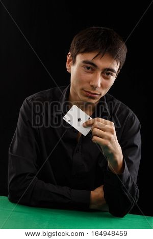 Beautiful young gambler man in black shirt sitting at the playing table with ace of spades in his hand isolated on black background