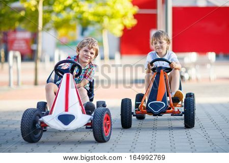 Two active little kid boys driving pedal race car in summer garden, outdoors. Children, best friends racing with fast speed and having fun. Twins making competition