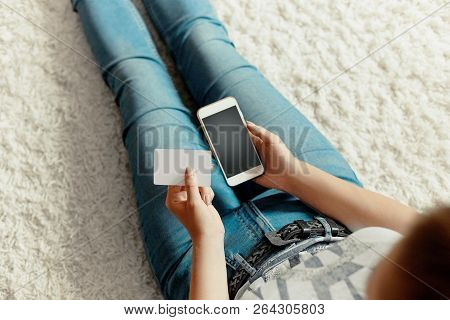 Young Woman Holding Credit Card And Using Phone Sitting On Floor. Payment.  Online Shopping Concept.