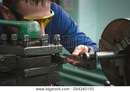 Turner Is Working On A Lathe Machine In A Factory. Worker Is Measuring A Produced Detail By Caliper.