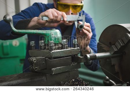 Turner Is Working On A Lathe Machine In A Factory. Worker Is Measuring A Produced Detail By Caliper.