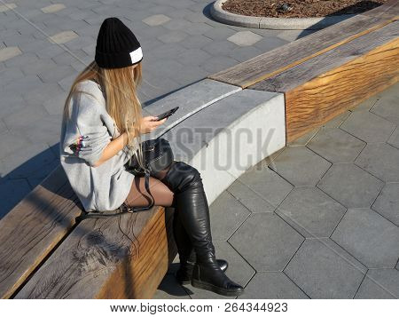 Teen Girl In A Black Cap And Jackboots Sitting With A Smartphone In Hands On The Street. Concept For