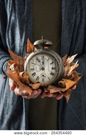 closeup of a young man with an old and rusty alarm clock surrounded by dry leaves in his hands, depicting the end of the summer time and the beginning of autumn