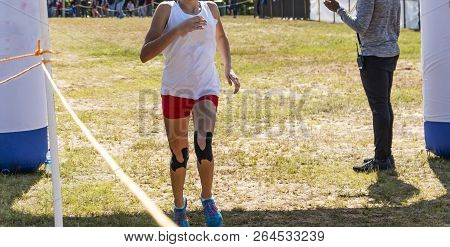 A Cross Country Runner With Black Tape On Her Knees Is Crossing The Finish Line In Frist Place.